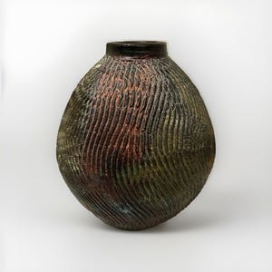 May include: A spherical ceramic vase with a narrow neck and a dark, textured surface. The exterior features deep, diagonal carved grooves with a mottled finish in shades of charcoal, deep green, and hints of copper-toned reddish-brown.