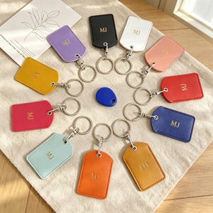 May include: A collection of rectangular keychains in various colors including black, white, pink, red, blue, orange, light blue, magenta, yellow, and purple. Each keychain features the letters MJ embossed in gold and a silver metal ring.