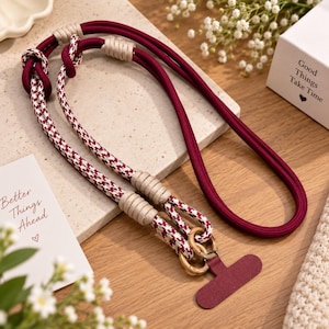 May include: A long phone lanyard featuring a solid deep burgundy cord and a patterned section with a white, burgundy, and black houndstooth design. It includes beige cord wrappings, two wooden rings, and a flat, T-shaped burgundy attachment.