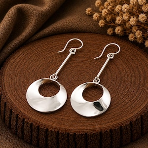 May include: A pair of silver-toned drop earrings featuring a polished, circular disc pendant suspended from a straight metal bar and a curved ear wire hook. The earrings have a smooth, reflective surface and a minimalist, geometric design.