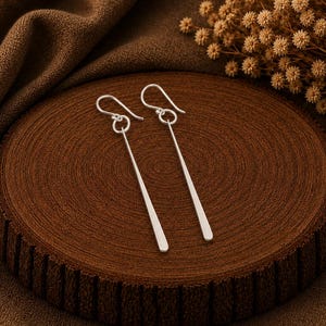 May include: A pair of silver-toned drop earrings featuring long, slender, flat bar pendants suspended from simple curved ear wires. The earrings have a smooth, polished finish and a clean, linear design that hangs vertically from the circular jump rings.