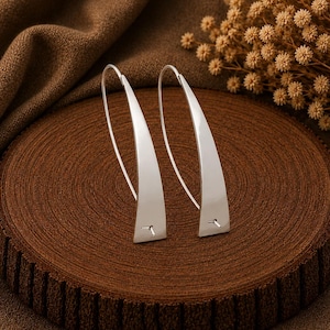 May include: A pair of silver-toned earrings featuring a curved, elongated triangular design. Each earring has a thin, arched wire hook that connects to the base of the smooth, polished metal form, creating a sleek, open-loop silhouette.