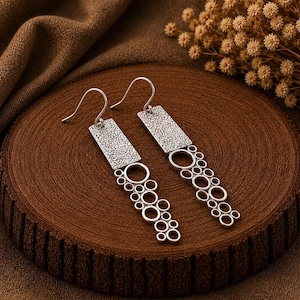May include: A pair of silver-toned drop earrings featuring a rectangular top section with a textured, hammered surface. Attached to the bottom of each rectangle is a cascading arrangement of open circular rings of varying sizes in a linear pattern.