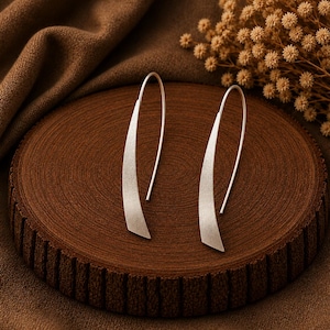 May include: A pair of silver-toned earrings featuring a long, curved, and tapered design with a matte, textured finish. The earrings are constructed with a simple hook-style wire that extends from the top of the slender, blade-like metal piece.