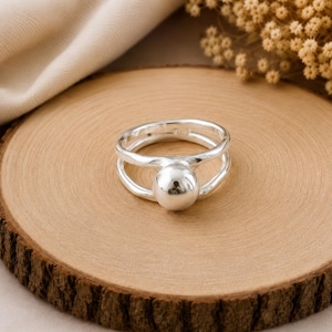 May include: A polished silver ring featuring a smooth, spherical ball accent centered on a double-band design. The ring is crafted from a reflective, metallic material with a continuous, rounded shape that connects the two bands at the front.