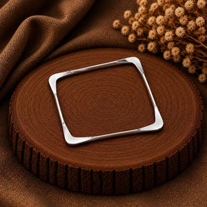 May include: A polished silver-toned bangle bracelet featuring a distinct square shape with rounded corners. The metal surface is smooth and reflective, showcasing a continuous, uniform band design that maintains a consistent thickness throughout its frame.
