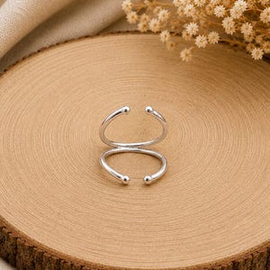 Pode incluir: Um anel aberto em tom prateado com um design de banda dupla. O anel consiste em dois fios curvos paralelos conectados de um lado, com cada uma das quatro extremidades finalizada com um pequeno detalhe liso e arredondado em forma de esfera.