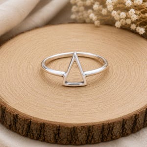 Pode incluir: Um anel de prata polida com uma forma geométrica triangular aberta única centrada na banda. O metal tem um acabamento liso e reflexivo, e o design consiste numa banda fina e contínua que forma um triângulo nítido no topo da peça.
