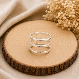 May include: A silver ring featuring a multi-band design with three distinct horizontal bands connected by a single vertical bar. The metal surface has a subtle hammered texture, creating a reflective finish across the circular structure.