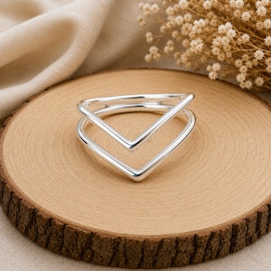 May include: A polished silver ring featuring a double V-shaped design. The band consists of two connected chevron-style segments that create a layered, geometric appearance. The metal surface is smooth, reflective, and has a bright metallic finish.