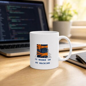 May include: A white ceramic mug featuring a graphic of a dark blue computer tower wrapped in orange tape and tangled wires, with the text "IT WORKS ON MY MACHINE" printed in blue capital letters below the illustration on the front side.