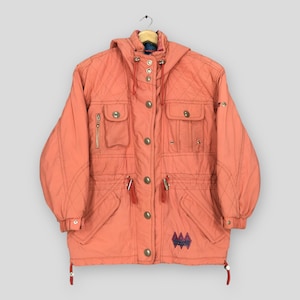 Könnte beinhalten: Eine korallenfarbene, gesteppte Jacke mit Kapuze, Frontreißverschluss mit Metalldruckknöpfen, mehreren Utility-Taschen an Brust und Taille sowie einem Kordelzug in der Taille. Ein kleines dunkelblaues und violettes geometrisches Logo ist unten.