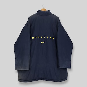 May include: A navy blue long-sleeve jacket with a high collar and snap buttons. The back features the word MICHIGAN printed in yellow capital letters above a small yellow Nike swoosh logo. The jacket has a solid dark blue color throughout.
