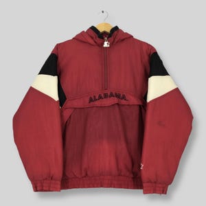 May include: A maroon pullover windbreaker jacket featuring a half-zip closure with a silver star zipper pull. The jacket has black and white color-blocked sleeves and the word ALABAMA embroidered in black across the front chest panel.