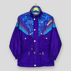 Könnte beinhalten: Eine violette Windjacke mit einem blau-violett gemusterten Oberteil, blaugrünen und violetten Chevron-Streifen und dem Wort BRENZO in gelber Schrift. Die Jacke verfügt über einen hohen Kragen, einen Frontreißverschluss und mehrere Taschen.