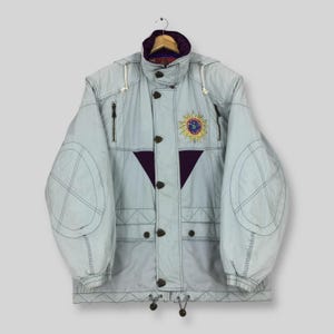 May include: A light blue hooded winter jacket featuring a high collar with a dark purple interior lining, a front zipper closure with dark buttons, and a yellow and blue sun-shaped embroidered patch on the chest with dark purple triangular accents.