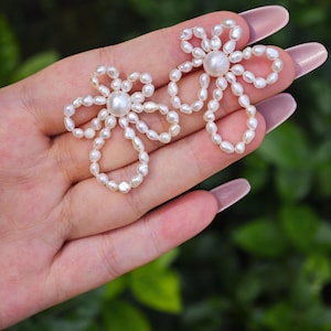 Pode incluir: Um par de brincos em forma de flor, feitos de pequenas pérolas de água doce brancas e irregulares. Cada brinco apresenta uma pérola redonda central rodeada por uma série de pétalas de pérolas em laço, criando um design floral aberto.
