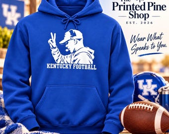 May include: A royal blue hooded sweatshirt featuring a white graphic of a person in a baseball cap making a peace sign, with the text Kentucky Football below. The Printed Pine Shop logo and the text Wear What Speaks to You are visible.