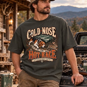 May include: A dark olive green short-sleeve t-shirt featuring a graphic print of a beagle dog chasing a rabbit in a mountain landscape. The text reads COLD NOSE HOT RACE and APPALACHIAN RABBIT HUNTING with a paw print icon at the bottom center.