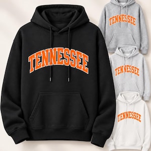 Op de afbeelding: Een zwarte hoodie met het woord TENNESSEE in een gebogen oranje lettertype met een witte omlijning, vergezeld van drie kleinere hoodies in grijs, lichtgrijs en wit, elk met hetzelfde oranje en witte tekstontwerp.