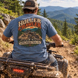 Peut inclure: Un t-shirt bleu à manches courtes avec un motif au dos portant le texte HOLLER CRAWLER et APPALACHIAN TRAIL CO. Le dessin représente un véhicule tout-terrain roulant dans une forêt avec des montagnes et un soleil en arrière-plan.