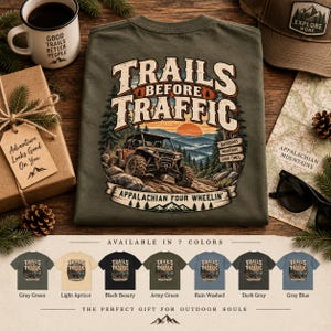 Op de afbeelding: Een olijfgroen t-shirt met een afbeelding van een terreinwagen op een bergpad, voorzien van de tekst Trails Before Traffic en Appalachian Four Wheelin'. Het ontwerp bevat dennenbomen, een zonsondergang en kleine houten bordjes.