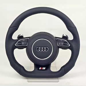 Op de afbeelding: Een zwart lederen stuurwiel met een geperforeerde grip, zilveren schakelflippers en geïntegreerde bedieningsknoppen op de spaken. De centrale naaf toont het Audi-logo in zilver, met een S5-embleem op de onderste spaak.