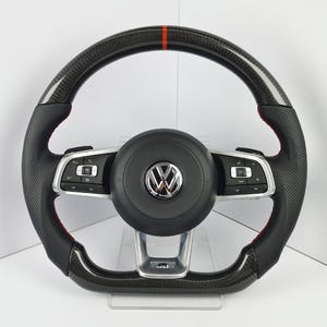 May include: A black Volkswagen steering wheel featuring a carbon fibre top and bottom rim with a red centre stripe. The side grips are made of perforated black leather with red stitching. It includes a central airbag cover with a chrome VW logo.