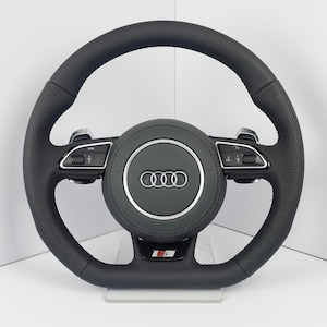 Op de afbeelding: Een zwart lederen Audi-stuurwiel met geperforeerde zijgrepen, zilveren schakelflippers en geïntegreerde bedieningsknoppen op de horizontale spaken. De centrale naaf toont het zilveren Audi-logo met vier ringen en een S-badge aan de onderkant.