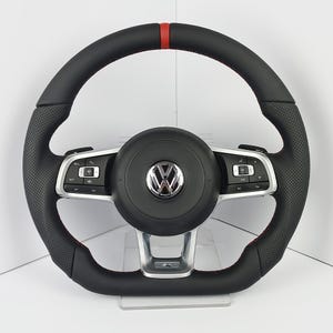 Puede incluir: Un volante de cuero negro con una franja roja en la parte superior, agarres laterales perforados y detalles metálicos plateados. El centro presenta un logotipo de Volkswagen cromado, flanqueado por botones de control en los radios.
