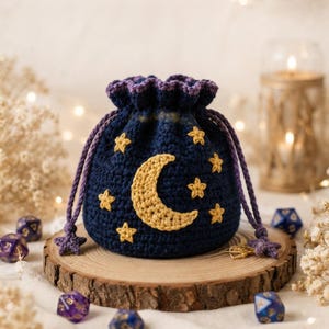 May include: A dark blue crochet drawstring pouch featuring a large yellow crescent moon and several small yellow stars embroidered on the front. The bag has a purple ruffled top edge and two purple braided drawstrings with star-shaped ends.