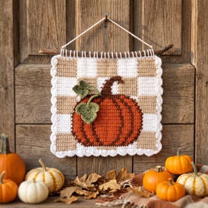 May include: A square crochet wall hanging featuring a white and tan checkered pattern with a large orange pumpkin and two green leaves in the center. The piece is suspended from a wooden branch by a white string and has a white scalloped border.