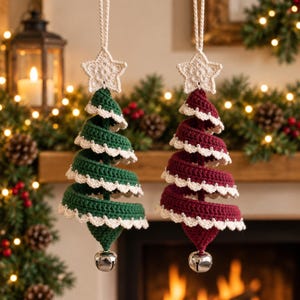 Op de afbeelding: Twee spiraalvormige kerstboomhangers van gehaakt garen, één in donkergroen en één in dieprood. Elke boom heeft een witte gehaakte ster aan de bovenkant, een witte geschulpte rand langs de zijkanten en een zilveren belletje aan de onderkant.