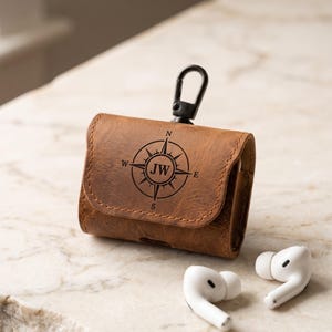 May include: A brown leather protective case for wireless earbuds featuring a black compass graphic with the letters JW in the center. The case includes a black metal carabiner clip attached to the top. Two white wireless earbuds are placed nearby.