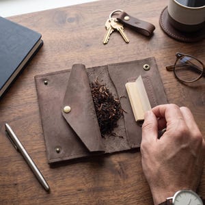 Pode incluir: Uma bolsa de tabaco em couro castanho com fecho de botão de pressão, contendo tabaco solto e um pequeno livrete de papel de enrolar. A bolsa está aberta, revelando uma superfície interior texturizada e detalhes metálicos na aba exterior.