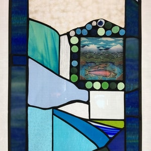 May include: A stained glass window with a blue, green, and white abstract design. A small rectangular panel in the center depicts a mountain scene with two salmon swimming in a river.