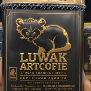 May include: A rectangular black metal coffee tin featuring a gold embossed civet cat illustration. The text reads LUWAK ARTCOFIE, LUWAK ARABICA COFFEE, KOPI LUWAK ARABIKA, and Nett weight 100g. It includes a Halal logo and production details.