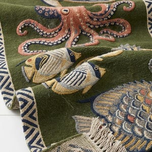 May include: A deep olive green rug featuring a textured, raised-pile design of various marine life, including an octopus and several fish in shades of blue, yellow, and coral. The rug has a geometric patterned border and a fringed edge at the bottom.