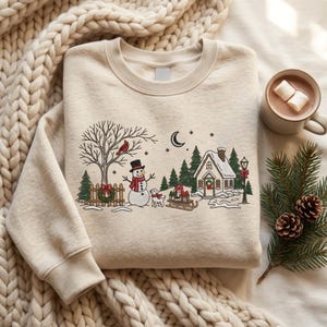 May include: A beige crewneck sweatshirt featuring an embroidered winter scene with a snowman, a dog, a house, a tree with a cardinal, a fence with a wreath, a sled with gifts, a lamppost, a crescent moon, and stars in black, red, green, and white.