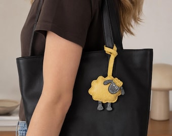 May include: A black tote bag featuring a yellow sheep-shaped bag charm attached to the handle. The sheep charm has a textured yellow body, a dark gray face with white eyes, and dark gray legs, hanging from a yellow strap tied to the bag handle.