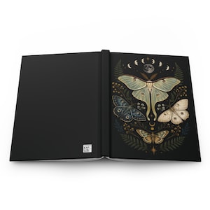 May include: A black hardcover journal featuring a wrap-around design. The back cover is solid black with a small white QR code. The front cover displays a detailed illustration of four moths, fern leaves, and a lunar cycle against a black background.