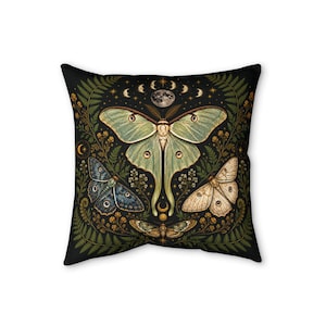 May include: A square throw pillow featuring a black background with a large central pale green moth, a smaller blue moth, a cream-colored moth, and a brown moth. The design includes green fern fronds, small yellow flowers, stars, and moon phases.