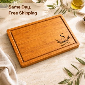 May include: A rectangular bamboo cutting board with a grooved perimeter and a natural wood grain finish. The board features a laser-engraved design consisting of a large letter S centered above the name SOPHIA, framed by two leafy olive branches.