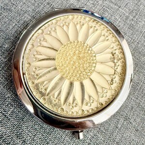 May include: A round, silver-toned compact mirror featuring a decorative top cover with a raised, cream-colored sunflower design. The center of the flower is textured with small, circular beads, surrounded by smooth, elongated petals and a border of dots.