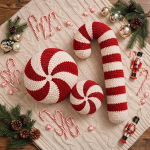 May include: A large crochet candy cane pillow and two round peppermint swirl pillows in red and white, surrounded by small plastic candy canes, silver ornaments, pine branches, and two nutcracker figurines on a cream-colored cable knit fabric.