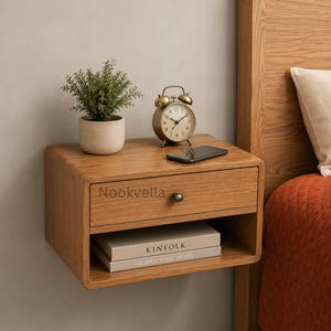 May include: A wall-mounted wooden nightstand with a single drawer featuring a round knob and an open shelf below. The surface holds a small potted green plant, a round analog alarm clock, and a black smartphone. The drawer is labeled Nookvella.