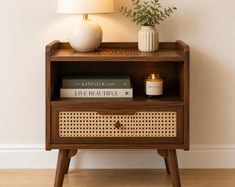 May include: A dark wood nightstand with four tapered legs, an open shelf, and a single drawer featuring a woven cane front panel. The nightstand holds a cream-colored table lamp, a small potted plant, two stacked books, and a glass candle jar.