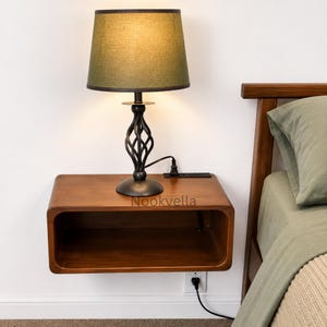 May include: A floating wooden nightstand with a smooth, rounded rectangular shape and a dark brown finish. On top sits a table lamp with a dark olive green fabric shade and a black metal base featuring a twisted, cage-like design. Text reads Nookvella.
