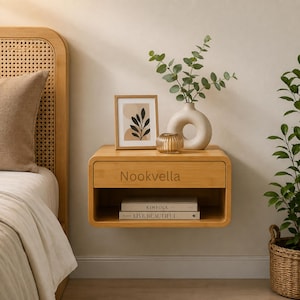 May include: A light wood floating nightstand with rounded corners and the text Nookvella on the drawer front. The unit features an open storage shelf containing two books, Kinfolk and Live Beautiful, and a top surface holding a picture frame, a vase, and a candle.