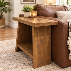 May include: A rectangular wooden side table with a natural brown wood grain finish, featuring a flat top surface and a lower shelf. The word Nookvella is printed on the top surface. The table is positioned next to a brown leather sofa.